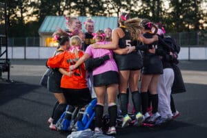 Field hockey team in a group huddle, wearing pink and black uniforms with bows, showing team spirit.