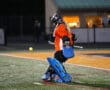 Field hockey goalie in action, wearing orange and blue gear, focuses on incoming ball on a turf field.