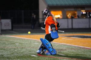 Field hockey goalie in action, wearing orange and blue gear, focuses on incoming ball on a turf field.