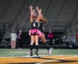 Two field hockey players in black jerseys and pink tutus celebrating with a jump on the field.