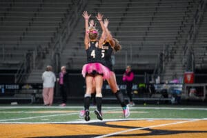 Two field hockey players in black jerseys and pink tutus celebrating with a jump on the field.