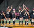Field hockey players in black uniforms celebrate with high-fives on a brightly lit field.