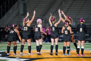 Field hockey players in black uniforms celebrate with high-fives on a brightly lit field.