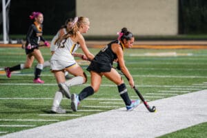 Youth field hockey game action with players in motion, girl in black and pink driving the ball down the field.