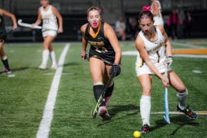 Two female field hockey players compete at night on a green field.
