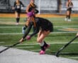 Field hockey player with purple hair dribbling the ball during a match on a green turf field.