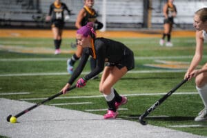 Field hockey player with purple hair dribbling the ball during a match on a green turf field.
