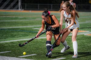Two female field hockey players compete for the ball on a turf field.