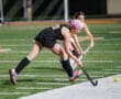 Two field hockey players in action on a turf field, competing for a yellow ball near the sideline.