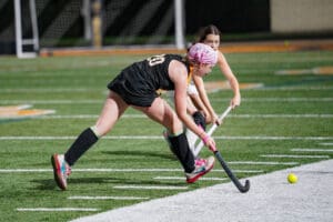 Two field hockey players in action on a turf field, competing for a yellow ball near the sideline.