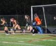 Field hockey game with players in black jerseys and goalie in orange, defending the goal at night.
