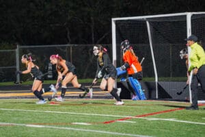 Field hockey game with players in black jerseys and goalie in orange, defending the goal at night.