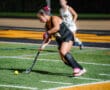 Field hockey player in black uniform dribbling ball on turf during a night game.