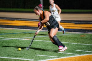 Field hockey player in black uniform dribbling ball on turf during a night game.