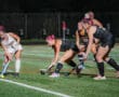 Field hockey game at night with players competing for the ball on a turf field.