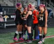 High school girls' field hockey team celebrating on the field in matching uniforms and pink bows.
