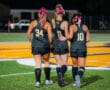 Three female field hockey players with pink ribbons walk together on the field wearing black uniforms.