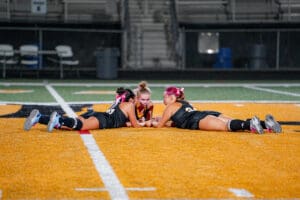 Three female athletes lie on a sports field, engaged in a focused discussion or game strategy session.