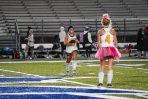 Two field hockey players in fun outfits warm up on a field, one wearing a pink tutu.