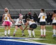 Field hockey team prepares on the field, some players wearing pink accessories for breast cancer awareness.