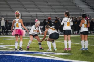 Field hockey team prepares on the field, some players wearing pink accessories for breast cancer awareness.