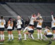 Girls' field hockey team celebrating victory on field under stadium lights.