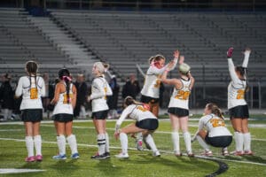 Girls' field hockey team celebrating victory on field under stadium lights.