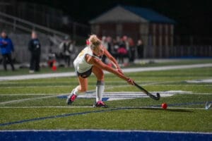 Field hockey player in action, striking the ball under stadium lights on the green turf.