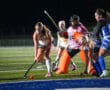 Field hockey match action with players competing for the ball under bright stadium lights.