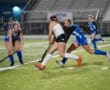 Field hockey players in action during a competitive match on a grassy field at night.
