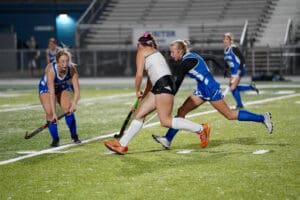 Field hockey players in action during a competitive match on a grassy field at night.
