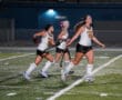 Teen field hockey players in action on a lit turf field at night, wearing white jerseys with Tigers text.