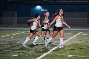 Teen field hockey players in action on a lit turf field at night, wearing white jerseys with Tigers text.