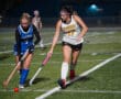 Two field hockey players from opposing teams in action on the field at night.