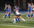 Girls playing a competitive field hockey match on a well-lit sports field, wearing blue and white uniforms.