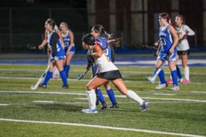Girls playing a competitive field hockey match on a well-lit sports field, wearing blue and white uniforms.