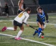Two female field hockey players compete for the ball on a grassy field during a game at night.