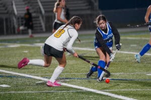 Two female field hockey players compete for the ball on a grassy field during a game at night.