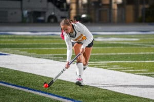 Field hockey player in action on a turf field, focused on the ball, wearing a white uniform and pink hair ties.
