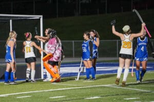 Girls' field hockey teams celebrating and discussing near goalpost on field.
