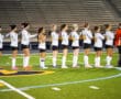 Girls' field hockey team lined up on field turf before a game, hands on their hearts.