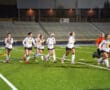 Girls' field hockey team running on the field, wearing white jerseys, preparing for a night game.