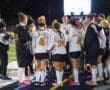 Women's field hockey team in a huddle on the field at night, preparing for a game.
