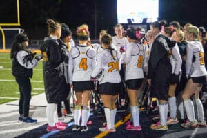 Women's field hockey team in a huddle on the field at night, preparing for a game.