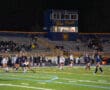 High school girls' field hockey game under stadium lights, with players in action and spectators in the stands.