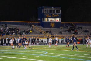 High school girls' field hockey game under stadium lights, with players in action and spectators in the stands.