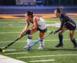 Two field hockey players compete for the ball on a green turf field during a match.