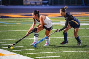 Two field hockey players compete for the ball on a green turf field during a match.