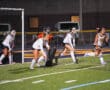 Field hockey players in action during a night game, focused on a defensive play near the goal.