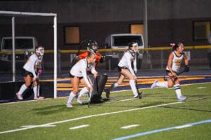 Field hockey players in action during a night game, focused on a defensive play near the goal.